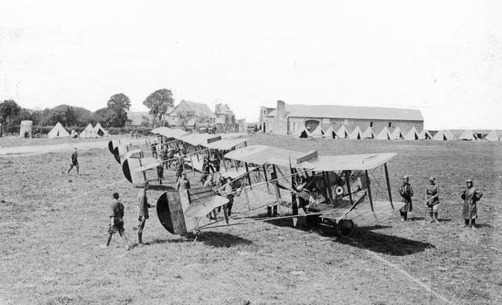 Period black and white photograph of a line of DH2 pusher scouts drawn up on a grass aerodrome, with bell tents, a hangar and airmen among the aircraft.